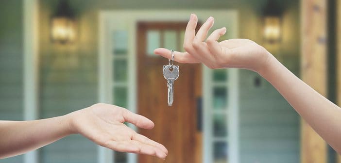 closeup of human hand with key dropping key into another person's open palm in front of a house