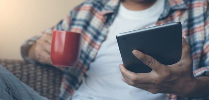 Casual man or hipster drinking coffee and looking at digital tablet screen near the window at home with morning light. Man checking internet application on digital tablet computer, modern lifestyle concept, flare light