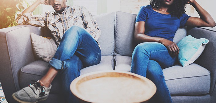 Young displeased black couple.American african men arguing with his girlfriend,who is sitting on sofa on couch next to him with legs crossed.Man looking away offended expression on her face.