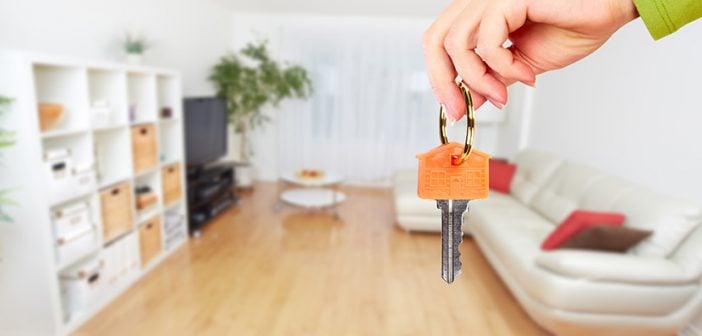 Hand with house key in foreground, living room with light wood floors, white couch and white shelves in background