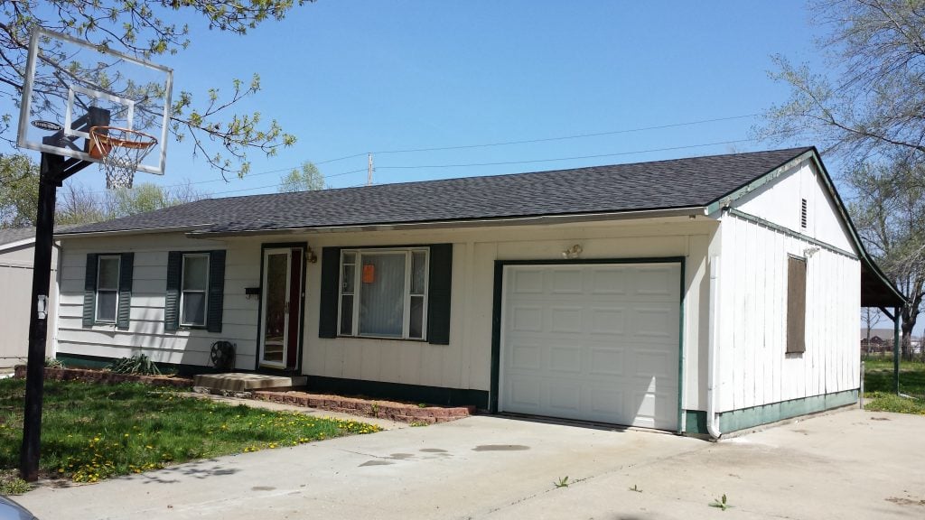 disheveled single-level white home with green trim and one-car garage with basketball hoop in driveway