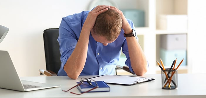 seated man looking down with hands on back of head and elbows on desktop worried, sad or frustrated at work