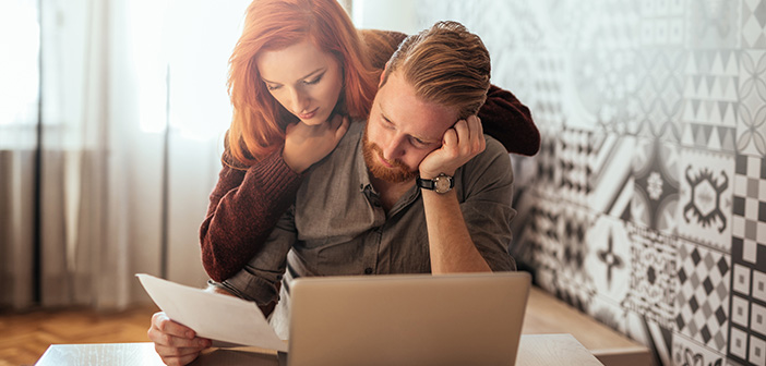 tired, frustrated young couple going over bill man sitting at desk with laptop woman looking over his shoulder