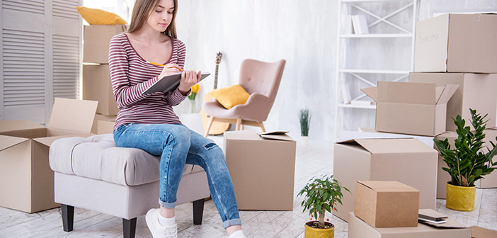 woman sitting amongst moving boxes in brightly lit living room jotting notes on a clipboard