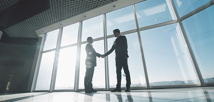 Two young businessmen are shaking hands with each other standing against panoramic windows.