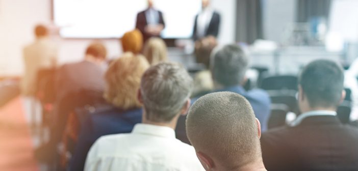 several people seated in a classroom setting and two suited male speakers at the front of the room