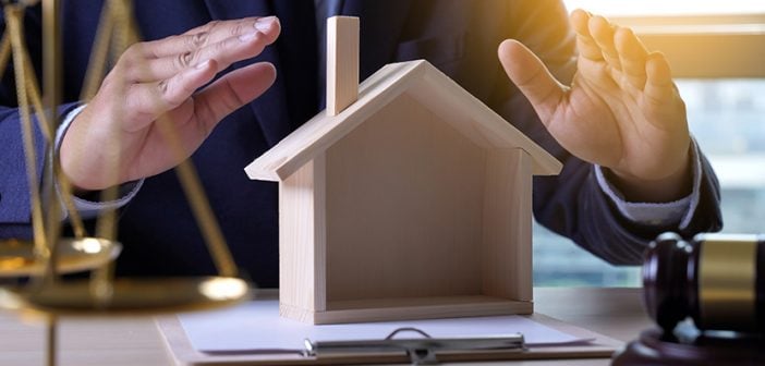 man in suit sitting at desk sheltering wooden model home on desk with hands, gavel and scale of justice in view