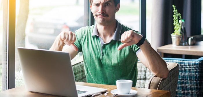 Dislike! Young dissatisfied businessman in green t-shirt sitting, working on laptop, looking at camera and showing thumbs down. business and freelancing concept. indoor shot near big window at daytime