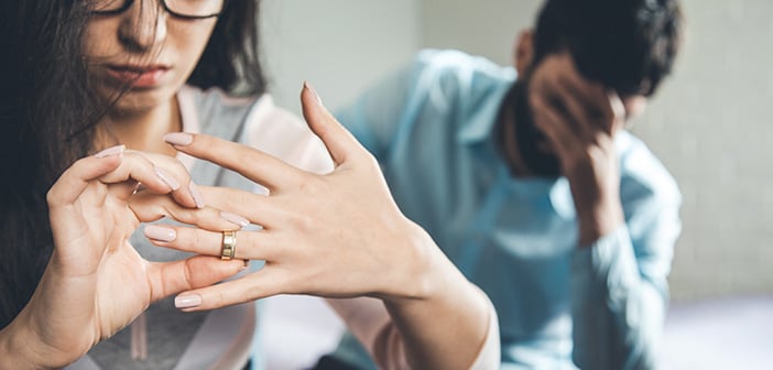 closeup of woman removing wedding ring from hand with man in background covering his face with his hand in despair