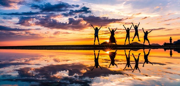 young people silhouettes jumping with sunset in background and water in foreground reflections of silhouettes clearly visible in water