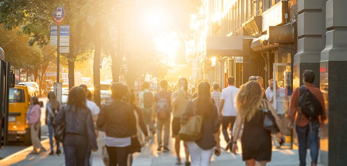 Man stands in the middle of a busy sidewalk looking at his cell phone while crowds of people walk around on 14th Street in Manhattan, New York City with the glow of sunlight in the background.