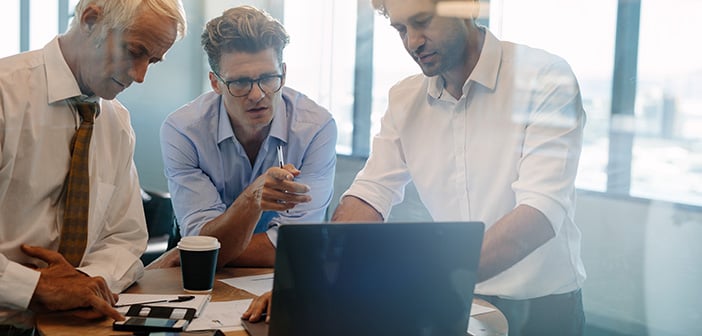 Business people talking while standing by table in office. Businessman giving demonstrating on laptop to colleagues