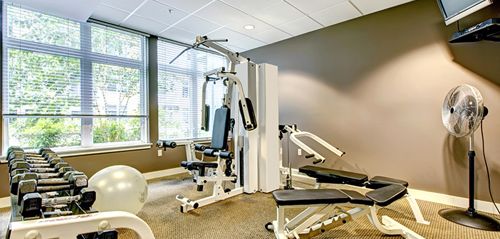 Gym in apartment building with brown wall, mounted TV, weights, benches, fan, exercise machines.