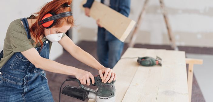 young woman sanding planks of wood with an orbital sander in an unfinished room during DIY renovations