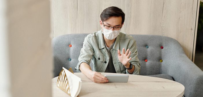 man seated at table wearing mask looking at phone in hand and waving to someone on video call