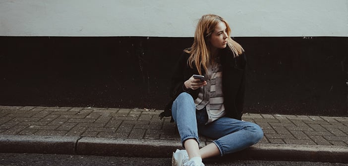 young woman waits on curb for ride seated with cell phone in hand