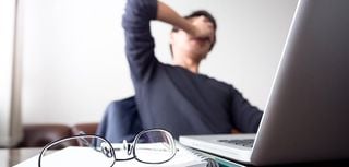 man at desk with hand covering face taking a break from work with glasses and open laptop on desk symbolizing stress, frustration