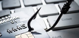 fishing hook pierced through credit card with blurred keyboard in background implying phishing scam