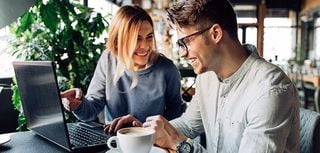 Attractive man in eyeglasses and charming woman is pointing at the laptop screen, laughing together, resting at cafe with cup of coffee.