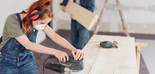 young woman sanding planks of wood with an orbital sander in an unfinished room during DIY renovations