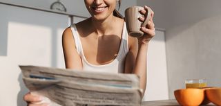 Smiling young girl standing at the kitchen in the morning, reading newspaper, drinking tea