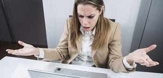 woman in business attired seated at desk looking at laptop computer with arms raised in wonderment and disappointment